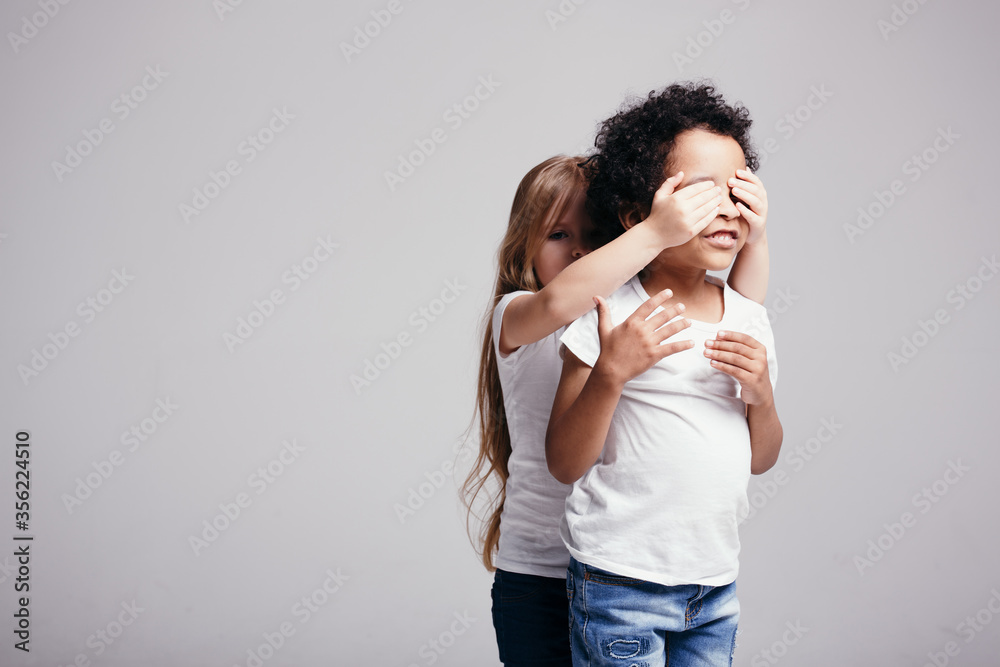The little blonde girl closed her curly dark-skinned boy's eyes so that he would guess who was behind him. The little blonde girl closed her curly dark-skinned boy's eyes so that he would guess who was behind him.