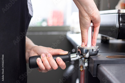 close-up photo of female hands holding a metal tamper and a portafilter with coffee in a coffee shop. A barista preparing for pressing ground coffee for brewing espresso or americano in a cafe