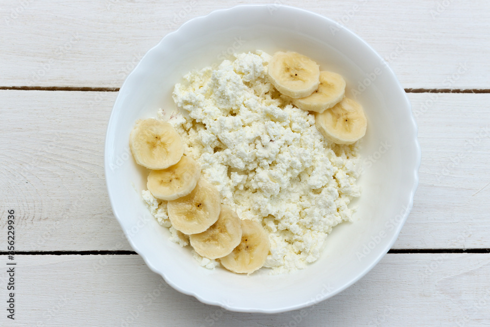Healthy breakfast. Cottage cheese in white bowl with banana on wood background