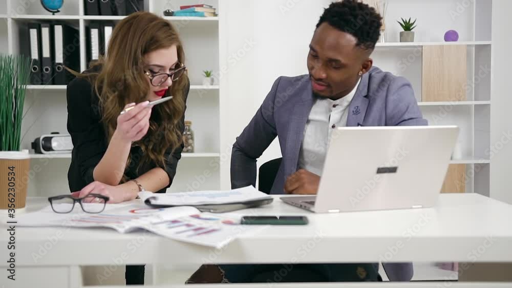 Front view of likable confident high-skilled businesswoman together with her african american bearded colleague which analyzing datas from report and datas from laptop in office Front view of likable confident high-skilled businesswoman together with her african american bearded colleague which analyzing datas from report and datas from laptop in office
