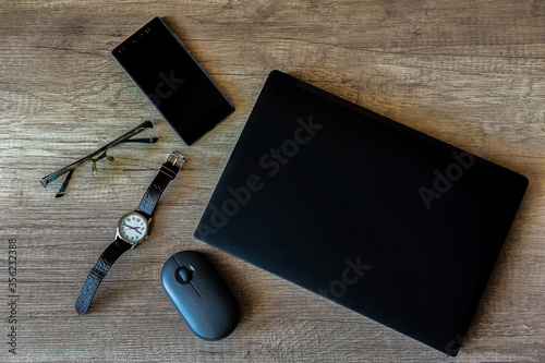 On a textured brown wooden table is a home work space with a black laptop, mobile phone, wristwatch, glasses, and computer mouse. Close-up view from above.