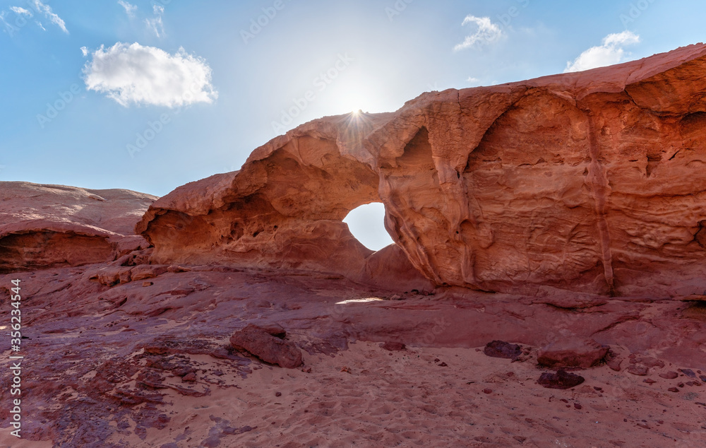Little arc or small rock window formation in Wadi Rum desert, bright sun shines on red dust and rocks, blue sky above
