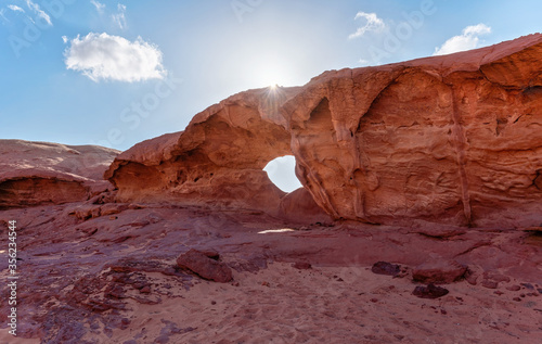 Little arc or small rock window formation in Wadi Rum desert, bright sun shines on red dust and rocks, blue sky above
