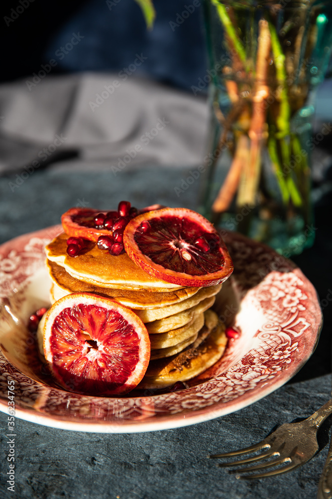 Vintage plate with pancakes with blood orange slices, red sauce and pomegranate seeds on dark background.
