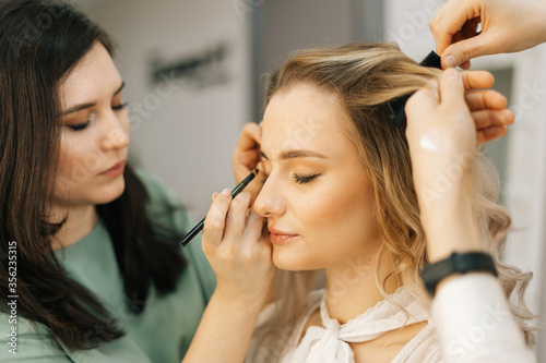Young pretty woman sitting in chair and getting professional make-up service from makeup artist and hairdresser in salon. Professional make-up artist works. Concept of backstage work.