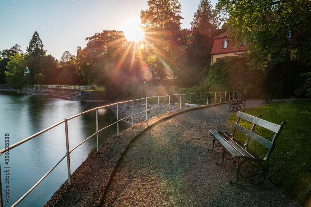 Obraz premium D, Bodensee, Lindau, Lindenhofpark, Bad Schachen, sonnenuntergang am Ufer mit leerer Sitzbank und Sonnenstern und glatter Wasserfläche (soft, warm)
