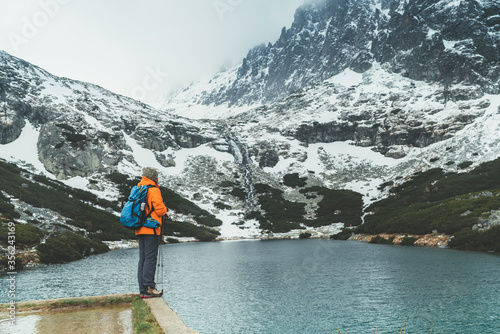 Dressed bright orange jacket female backpacker enjoying the Velicke pleso (mountain lake) view as she have mountain walk in Velicka valley in High Tatras, Slovakia. Active people in nature concept.