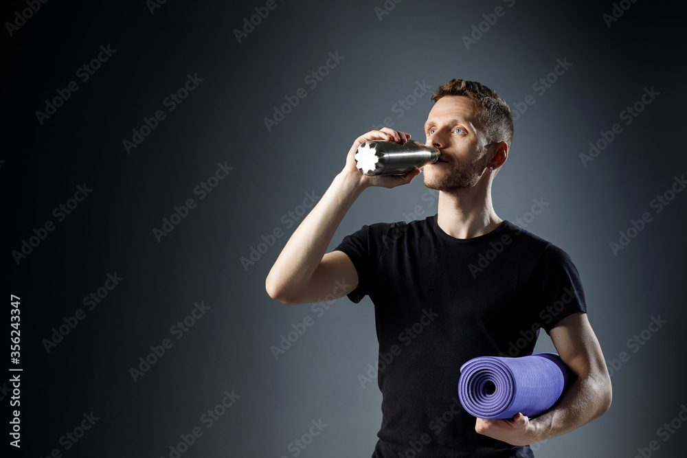 Young man on the black background is drinking water from fitness bottle, holding fitness mat.