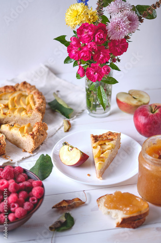 A sliced rustic apple pie on a linen towel. Dessert on a white wooden table, around are apples, raspberries in a plate, apple jam and flowers.