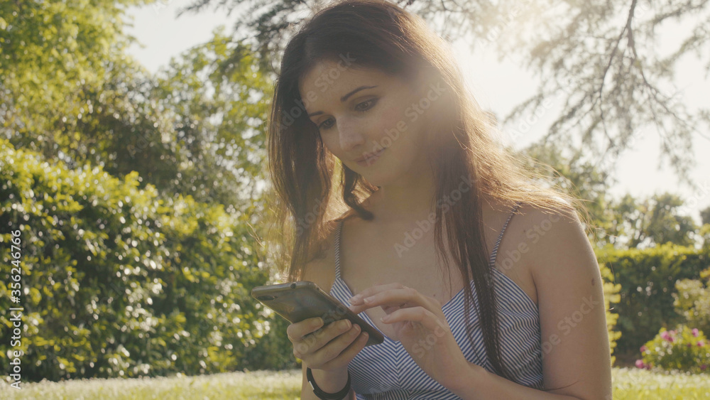 Fototapeta premium Cute young woman holding mobile phone in hand typing browsing online on social media, relax rest outdoor in home garden grass, sunny day in spring summer season. blurred background