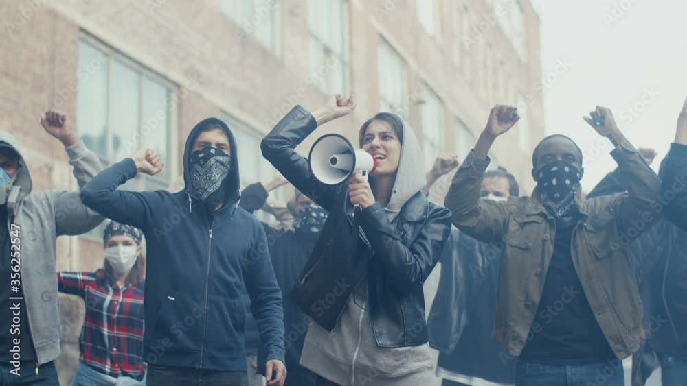 Caucasian young woman in hood screaming mottos in megaphone. Multiethnic male and female crowd of protesters shouting slogans and protesting. Girl leading at manifestation for human rights.