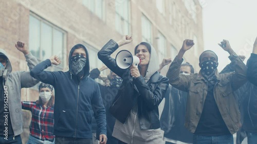 Caucasian young woman in hood screaming mottos in megaphone. Multiethnic male and female crowd of protesters shouting slogans and protesting. Girl leading at manifestation for human rights. photo