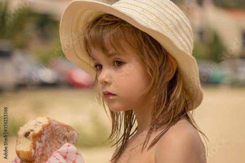 Beautiful little girl with hat eating a doughnut on the beach.