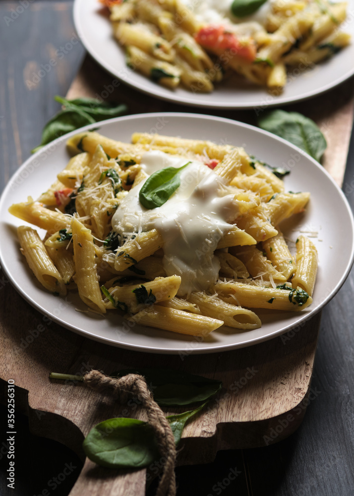 Two portions of pasta with tomatoes, spinach, parmesan and bolognese sauce on a large kitchen wooden board. Dish in a plate on a wooden table.