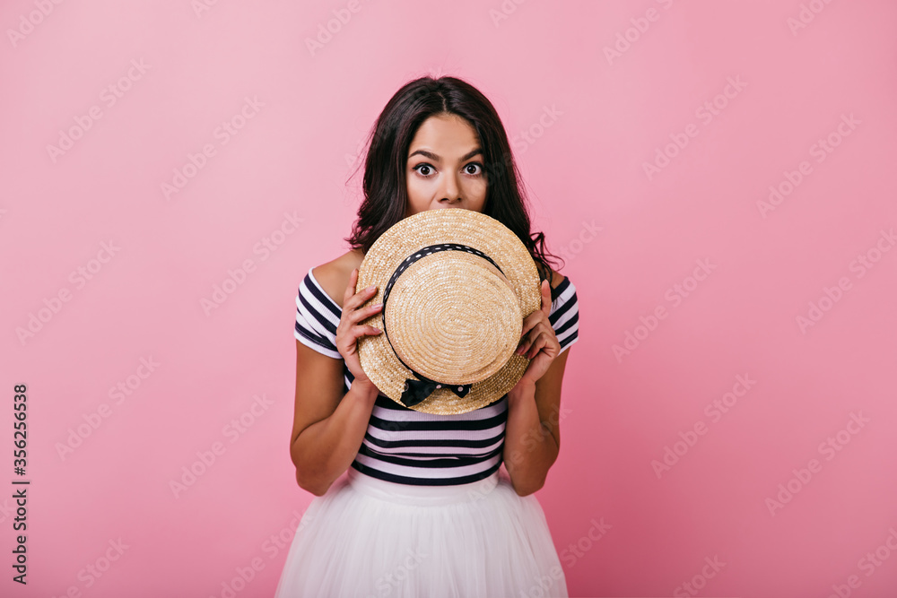 Graceful tanned woman hiding face beside straw hat. Indoor portrait of adorable latin lady playfully posing on pink background.