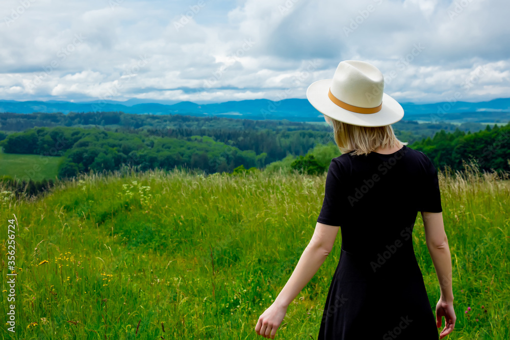 Blonde woman in black dress and white hat at meadow with mountains on background Blonde woman in black dress and white hat at meadow with mountains on background