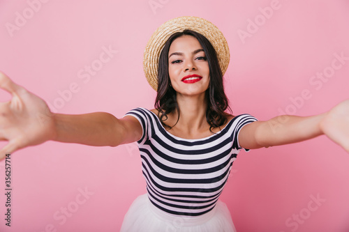 Charming young woman with black eyes making selfie on pastel background. Indoor photo of refined glamorous girl having fun in studio.