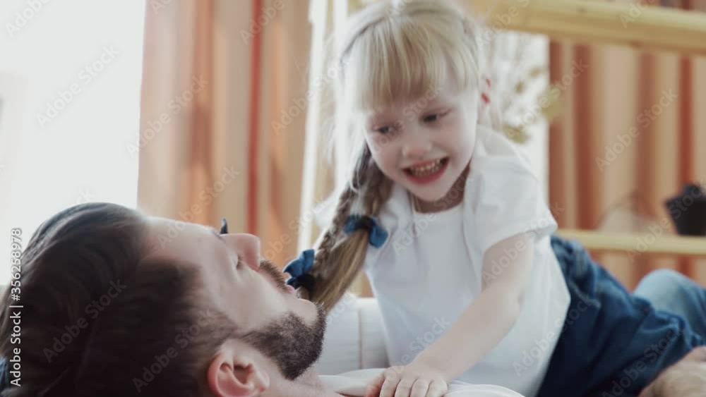 Close up of young man in jeans and white t-shirt plays with his daughter on couch Close up of young man in jeans and white t-shirt plays with his daughter on couch
