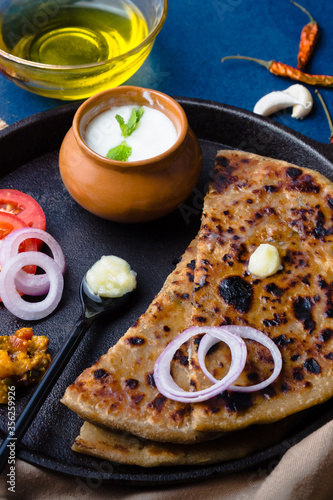 Aloo Paratha / Indian Potato Stuffed Wheat Bread served with Ghee (Indian Butter), Curd & Pickle, Tomato & Onion. Top View on a Dark Moody Background. Traditional North Indian or Punjabi Breakfast.