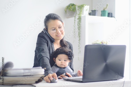 Madre feliz trabajando en la computadora desde su casa y hablando por teléfono mientras sostiene a su bebé. Teletrabajo en la cuarentena.