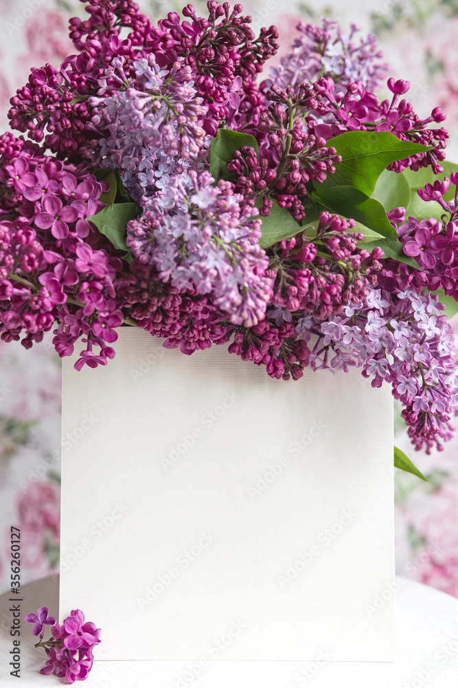 White sheet in beautiful blooming lilac on a white stand on a floral background. Greeting card, place for text, mock up