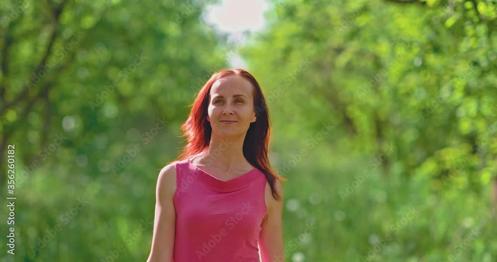 Beautiful young woman walks in the spring garden and touches the branches of green trees. Woman is happy, she is smiling. Walk on a spring day in a tree garden with green foliage.