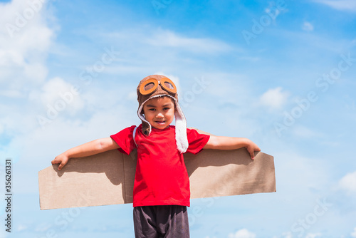 Happy Asian funny child or kid little boy smile wear pilot hat play and goggles with toy cardboard airplane wing flying outdoor against summer blue sky cloud background, Startup freedom concept