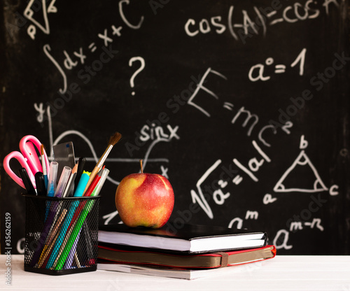 Coloured school supplies, apple, books on white contertop on chalkboard background with formulas. Education concept