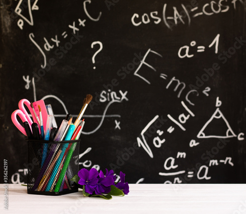 Coloured school supplies and flowers on white wooden contertop on chalkboard background with formulas. Education concept