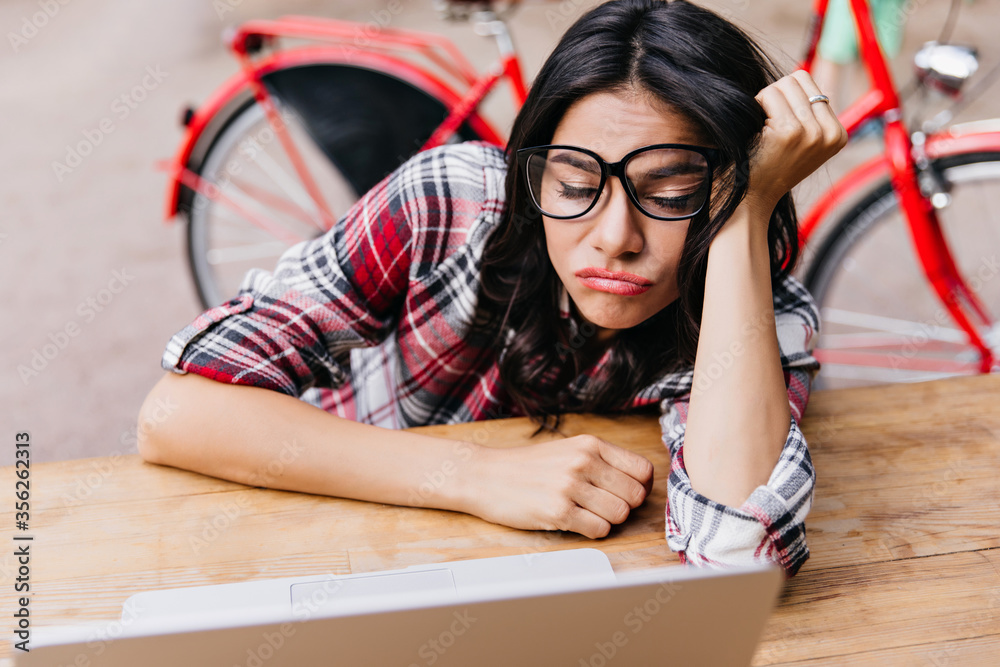 Tired female student posing on the street with bicycle on background. Outdoor portrait of sleeping brunette girl sitting near laptop. Tired female student posing on the street with bicycle on background. Outdoor portrait of sleeping brunette girl sitting near laptop.