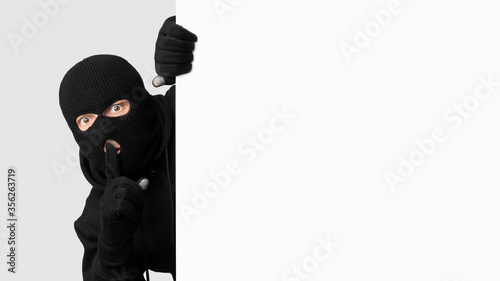 Photo Masked thief peeking out white blank board, making hush sign
