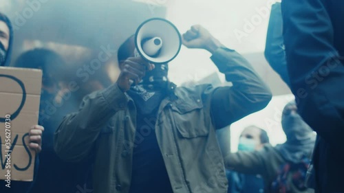 African American young handsome guy with scarf on face protesting in middle of crowd of protesters and screaming mottos in megaphone. Guy leading at manifestation and riot for human rights in smoke.