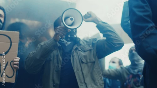 African American young handsome guy with scarf on face protesting in middle of crowd of protesters and screaming mottos in megaphone. Guy leading at manifestation and riot for human rights in smoke. photo