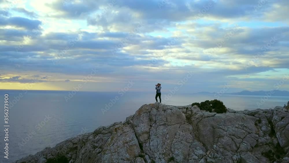AERIAL WS Young woman with binoculars standing on top of cliff of Cap Formentor at sunset/ Majorca, Spain AERIAL WS Young woman with binoculars standing on top of cliff of Cap Formentor at sunset/ Majorca, Spain