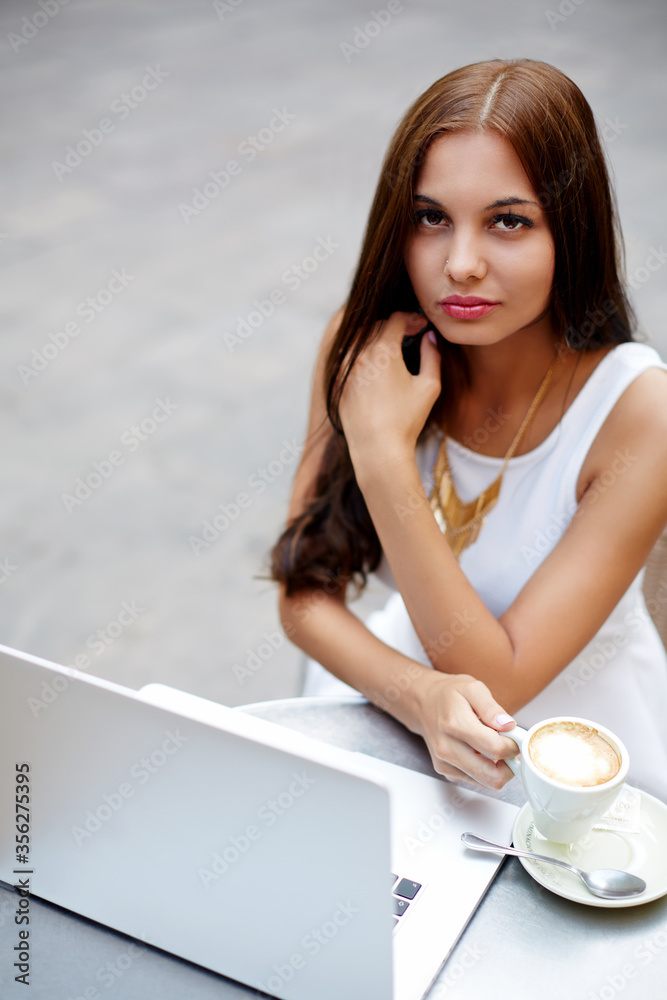 Young gorgeous woman drinking coffee while sitting with open laptop computer outdoors Young gorgeous woman drinking coffee while sitting with open laptop computer outdoors