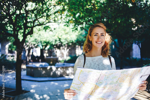 Young charming female tourist looking at camera and holding city map while standing in beautiful park