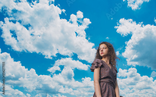 A slender, beautiful girl in a summer sundress against the background of a beautiful cloudy sky and a green river bank