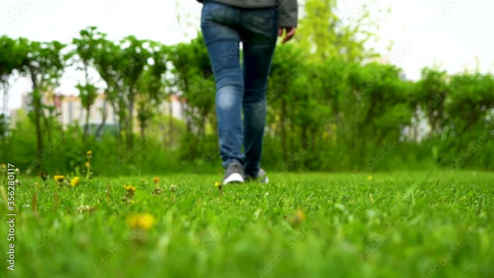 Footsteps on the grass are large with a blurred background. Close-up of a man's feet in sneakers and jeans walking on bright green grass in spring. Slow motion. Footsteps on the grass are large with a blurred background. Close-up of a man's feet in sneakers and jeans walking on bright green grass in spring. Slow motion.