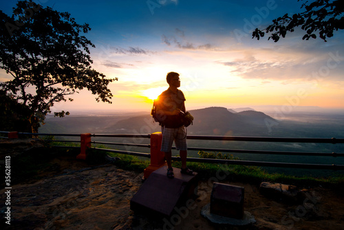 A photographer standing on top of Mountain at sunset with Mist.Asian male backpack in nature during sunset Relax time on holiday concept travel at Pha mor i dang,Sisaket province,Thailand,ASIA.