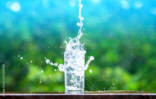 Drink water pouring in to glass over sunlight and natural green background.Water splash in glass Select focus blurred background.