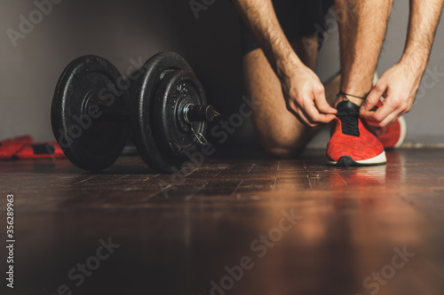 Morning workout routine in home gym. Fitness motivation and muscle training concept. Man in sneakers tying shoelaces. Athlete starting exercise with dubbell weight. photo