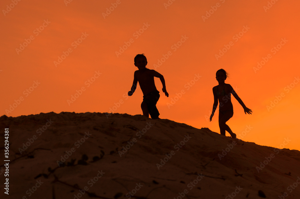 Siluetas de niños corriendo sobre duna con atardecer naranja durante vacaciones de verano.