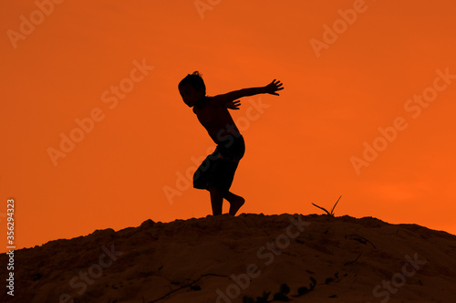 Silueta de niño a punto de saltar desde duna de playa con atardecer naranja en vacaciones de verano.