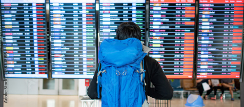 Vacation travel concept. Asian man backpacker and tourist carrying blue backpack checking flight information schedule on arrival departure board in international airport terminal.