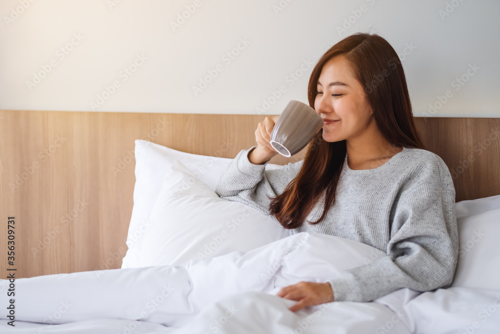 A beautiful asian woman drinking hot coffee while lying on a white cozy bed at home in the morning A beautiful asian woman drinking hot coffee while lying on a white cozy bed at home in the morning