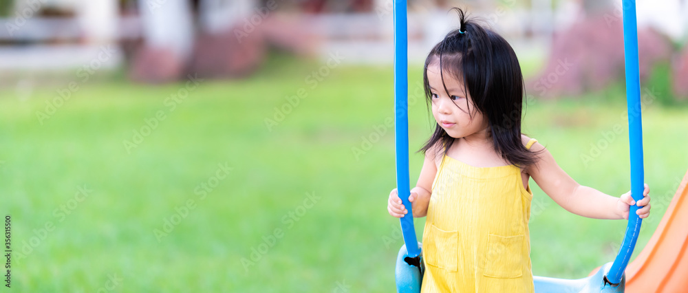 Happy Asian little child girl aged 3 years old is playing in a blue swing. Children wearing yellow jumpsuit, playing on the playground at home. The background is a green lawn. Widescreen photos.