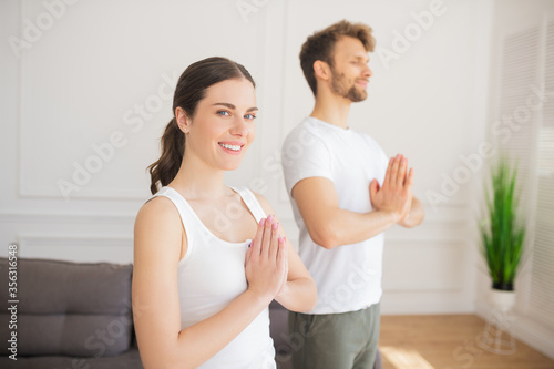 Young couple in white tshirts doing yoga at home putting their hands in namaste, woman smiling