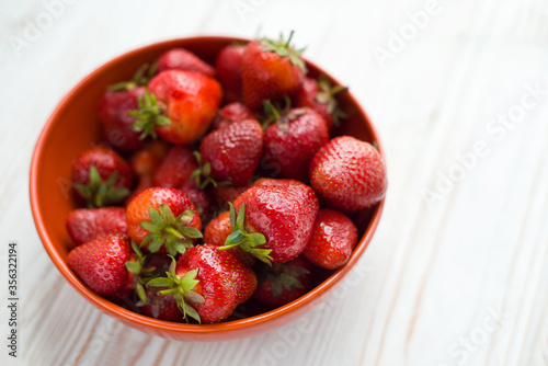 Close-up fresh ripe delicious strawberries in a orange bowl plate on white wooden table background. Antioxidant nutrition, healthy organic food concept. Copyspace. Flat lay. Daylight