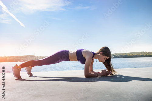 young female athlete doing push-ups outdoors near the lake at daytime. Healthy lifestyle.