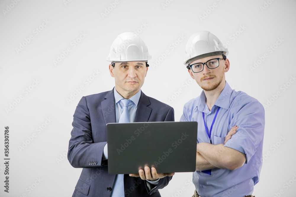 Serious engineer and architect in hard hats looks at camera, discuss new project while using laptop, making calculated engineering decisions isolated on white background. BIM industry construction Serious engineer and architect in hard hats looks at camera, discuss new project while using laptop, making calculated engineering decisions isolated on white background. BIM industry construction